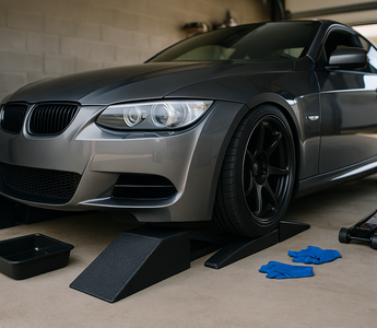 A lowered sports car positioned on split-style race ramps in a garage, with an oil drain pan and tools beside it