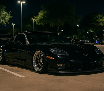 Slammed black Corvette on polished wheels parked at a nighttime car meet in a well-lit Texas parking lot.