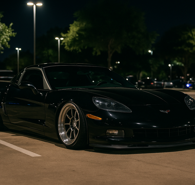 Slammed black Corvette on polished wheels parked at a nighttime car meet in a well-lit Texas parking lot.