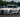 Widebody white BMW E36 coupe with angle kit andwith extended fenders and deep-dish wheels drifting at an outdoor motorsport event, captured mid-corner with spectators and tents in the background.