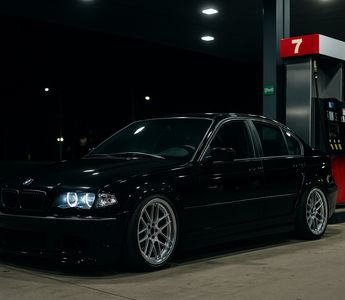 Lowered BMW E46 4-door on coilovers parked beside a gas station pump at night, showing aggressive stance and performance suspension setup.