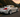 Slammed Infiniti G37S with wide body kit and huge rear wing rolling through a Japanese mountain road at sunset, captured mid corner with motion blur.