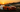 Drift car sliding through a corner in controlled oversteer, smoke rolling off rear tires on a racetrack during sunset