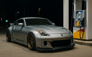 Slammed silver Nissan 350Z with a wide body kit parked at a gas station pump at night, illuminated by neon lights reflecting off wet pavement, showcasing aggressive stance and lowered coilover suspension.