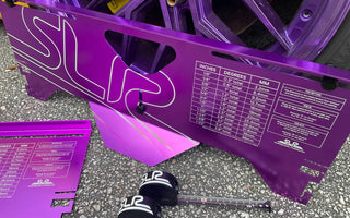 Purple aluminum toe plates leaning against a car wheel during alignment in a garage, with a tape measure showing toe measurement setup