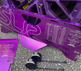 Purple aluminum toe plates leaning against a car wheel during alignment in a garage, with a tape measure showing toe measurement setup